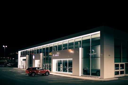 A car dealership at night with bright lights illuminating the building's modern, glass-front design. A red SUV is parked near the entrance, and the interior showcases more vehicles. The dealership is branded with the Volkswagen logo and the name St. John's is visible.
