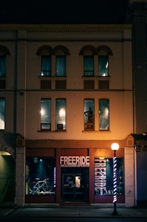 A storefront with a sign reading 'FREERIDE MOUNTAIN SPORTS' is lit by a streetlamp at night. The building has a vintage architectural style with arched windows on the upper floor. A bicycle is displayed in the window, and the interior lighting casts a warm glow.
