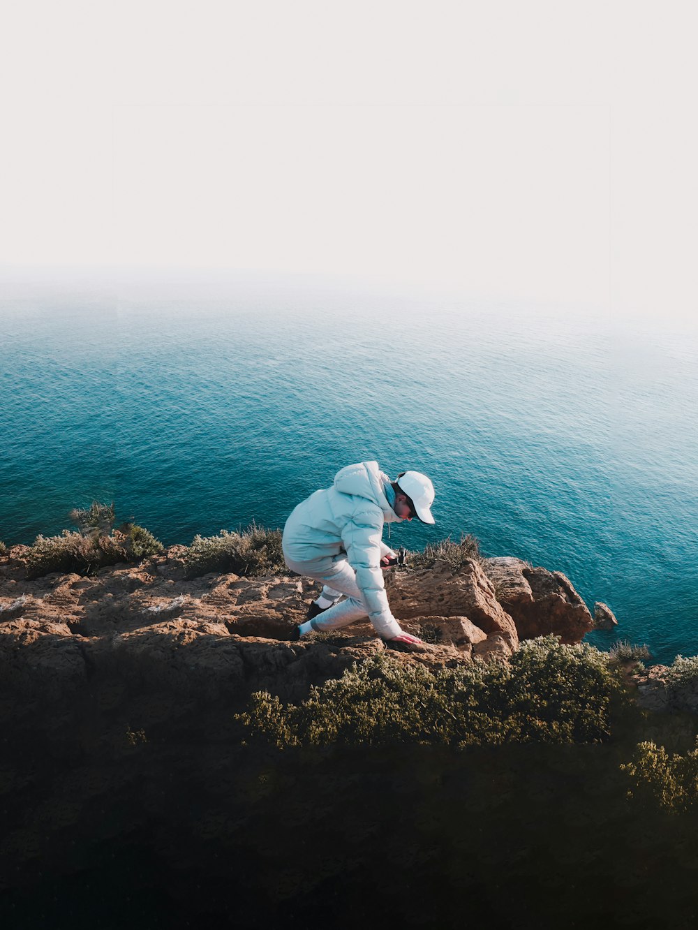 man in white long sleeve shirt sitting on brown rock formation near body of water during