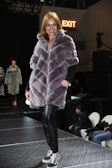 A woman wearing a plush, quilted, grey fur coat walks confidently down a runway. She is paired with black leather pants and black sneakers with white laces. The surroundings include a dimly lit stage with onlookers seated on the sides. A sign with 'EXIT' in red letters is visible in the background.