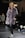 A woman wearing a plush, quilted, grey fur coat walks confidently down a runway. She is paired with black leather pants and black sneakers with white laces. The surroundings include a dimly lit stage with onlookers seated on the sides. A sign with 'EXIT' in red letters is visible in the background.