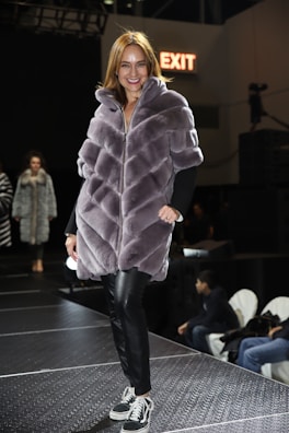 A woman wearing a plush, quilted, grey fur coat walks confidently down a runway. She is paired with black leather pants and black sneakers with white laces. The surroundings include a dimly lit stage with onlookers seated on the sides. A sign with 'EXIT' in red letters is visible in the background.