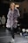 A woman wearing a plush, quilted, grey fur coat walks confidently down a runway. She is paired with black leather pants and black sneakers with white laces. The surroundings include a dimly lit stage with onlookers seated on the sides. A sign with 'EXIT' in red letters is visible in the background.