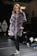 A woman wearing a plush, quilted, grey fur coat walks confidently down a runway. She is paired with black leather pants and black sneakers with white laces. The surroundings include a dimly lit stage with onlookers seated on the sides. A sign with 'EXIT' in red letters is visible in the background.