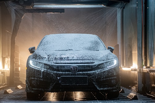 A black car is in the middle of a car wash. The vehicle is covered in soap foam and illuminated by the bright lights of the wash station. Water is spraying from multiple directions, creating a dynamic and dramatic effect as the car's headlights pierce through the suds.