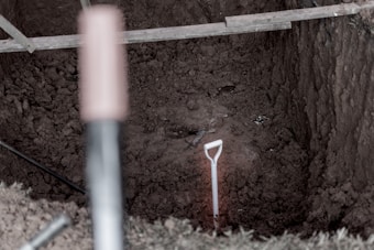 A shovel with a white handle is partially buried in a deep, freshly dug pit with earthy walls. The pit appears to be quite deep with scattered loose soil at the bottom. There are wooden planks laid across the top of the excavation, likely for support or safety. The foreground is blurred, featuring a partially shown object, adding depth to the scene.