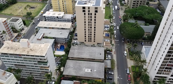 An aerial view of an urban area featuring several mid-rise residential and commercial buildings with flat roofs. The landscape includes a grid of streets lined with cars and palm trees. There's a mix of modern and older architectural styles, with green spaces and a small park visible in the background.