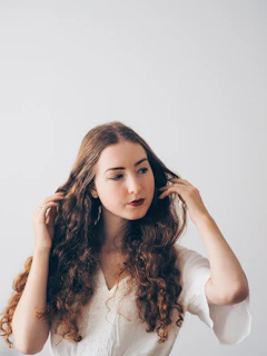 A serene close-up of a woman gently touching her glossy, healthy hair bathed in soft natural light with a muted sage green background.