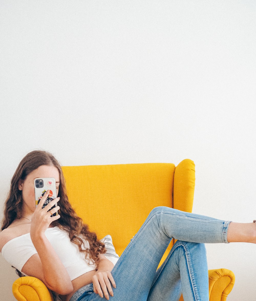 Frau in weißem Tanktop und blauer Jeans sitzt auf gelbem Sofa