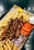 A serving of grilled meat skewers accompanied by French fries, tomato slices, sliced onions, pita bread, a lemon wedge, and a small bowl of white dipping sauce on a tray lined with paper.