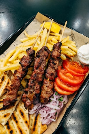 A serving of grilled meat skewers accompanied by French fries, tomato slices, sliced onions, pita bread, a lemon wedge, and a small bowl of white dipping sauce on a tray lined with paper.