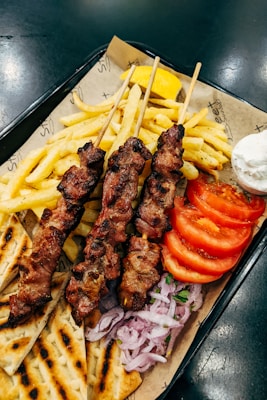 A serving of grilled meat skewers accompanied by French fries, tomato slices, sliced onions, pita bread, a lemon wedge, and a small bowl of white dipping sauce on a tray lined with paper.