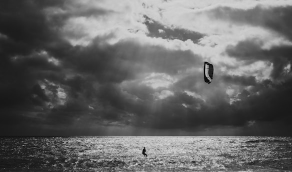 A dramatic black and white scene of a kitesurfer navigating the waves under a cloudy sky. The kite is prominently visible against the textured clouds, and sunlight filters through, creating a shimmering effect on the water.