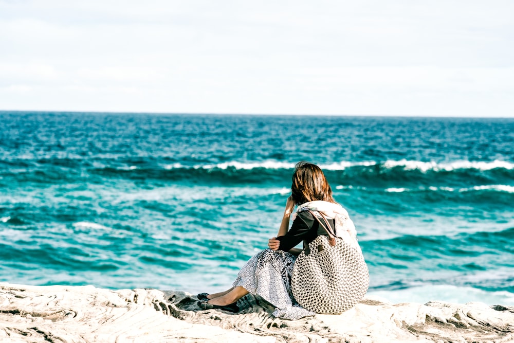 Frau in schwarz-weißem Polka-Dot-Kleid sitzt auf grauem Felsen in der Nähe von Wasser