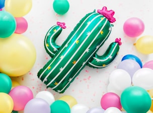 A vibrant scene with a green cactus-shaped balloon adorned with pink crowns, surrounded by a variety of colorful balloons including yellow, aqua, pink, and white. The background is light with small scattered pink decorations, conveying a festive atmosphere.