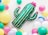 A vibrant scene with a green cactus-shaped balloon adorned with pink crowns, surrounded by a variety of colorful balloons including yellow, aqua, pink, and white. The background is light with small scattered pink decorations, conveying a festive atmosphere.