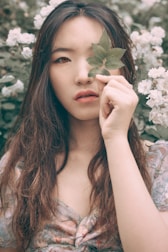 A woman with long hair stands among white flowers, holding a green leaf over one eye. The soft, natural light and floral background create a serene and artistic atmosphere.