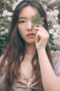 A woman with long hair stands among white flowers, holding a green leaf over one eye. The soft, natural light and floral background create a serene and artistic atmosphere.