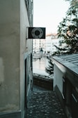 A narrow cobblestone alleyway between old buildings leads toward a river. The view includes a black sign with white text that reads 'SCHIPFE33 ZÜRICH 8001' mounted on one of the walls. In the background, a few trees and historic buildings are visible near the water, accompanied by a small street lamp.