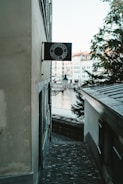 A narrow cobblestone alleyway between old buildings leads toward a river. The view includes a black sign with white text that reads 'SCHIPFE33 ZÜRICH 8001' mounted on one of the walls. In the background, a few trees and historic buildings are visible near the water, accompanied by a small street lamp.
