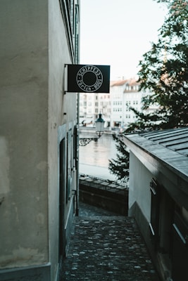 A narrow cobblestone alleyway between old buildings leads toward a river. The view includes a black sign with white text that reads 'SCHIPFE33 ZÜRICH 8001' mounted on one of the walls. In the background, a few trees and historic buildings are visible near the water, accompanied by a small street lamp.