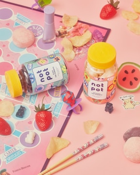 A playful and colorful arrangement featuring two jars labeled 'not pot' with quirky designs, surrounded by candy, potato chips, a strawberry, nail polish, chopsticks with cute patterns, and a game board. Various stickers, including a watermelon and cute figures, are scattered around, adding a whimsical touch.