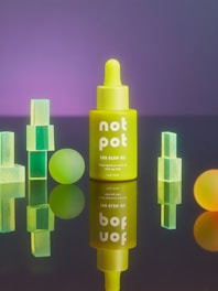 A neon yellow dropper bottle labeled 'not pot CBD Glow Oil' is centrally positioned, surrounded by geometrically shaped translucent objects including cubes and spheres. The background has a gradient of purple, adding a vibrant contrast to the foreground elements.