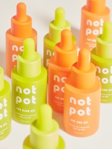 A collection of vibrant bottles with droppers in bright neon green and orange hues. Each bottle is labeled with 'not pot' and is part of a CBD product line, including glow oil and pet CBD oil.