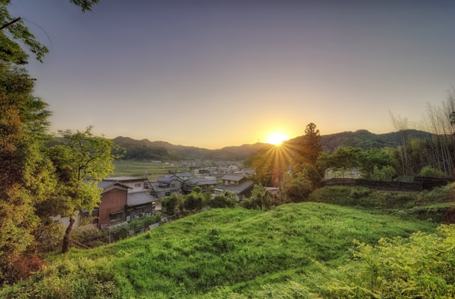 A serene rural landscape at dawn featuring a cluster of houses nestled among lush greenery, with the sun rising over distant hills. The foreground is dominated by vibrant green fields and trees, while the background transitions into gently rolling hills under a clear sky.