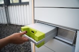 A hand with neatly manicured nails is placing or removing a metallic battery with yellow-green accents from a gray storage compartment. The surrounding environment is industrial, featuring a window with bars and a stone-covered ground.