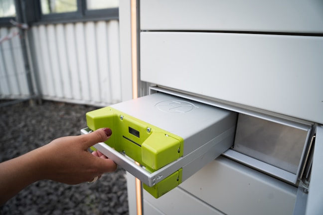 A hand with neatly manicured nails is placing or removing a metallic battery with yellow-green accents from a gray storage compartment. The surrounding environment is industrial, featuring a window with bars and a stone-covered ground.