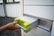 A hand with neatly manicured nails is placing or removing a metallic battery with yellow-green accents from a gray storage compartment. The surrounding environment is industrial, featuring a window with bars and a stone-covered ground.