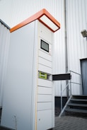 A tall, modern parking ticket machine with an orange top and digital display stands outside next to a corrugated metal building. It features multiple slots and compartments, with one section open to reveal internal components. The scene includes a set of metal stairs leading to a door in the background.