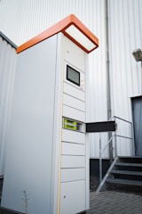 A tall, modern parking ticket machine with an orange top and digital display stands outside next to a corrugated metal building. It features multiple slots and compartments, with one section open to reveal internal components. The scene includes a set of metal stairs leading to a door in the background.