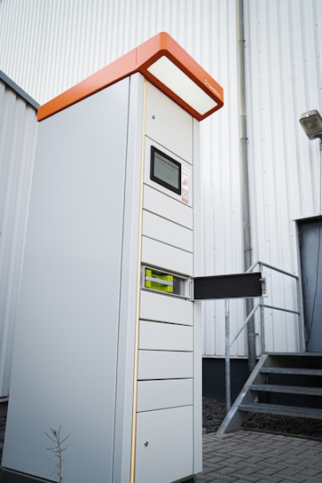 A tall, modern parking ticket machine with an orange top and digital display stands outside next to a corrugated metal building. It features multiple slots and compartments, with one section open to reveal internal components. The scene includes a set of metal stairs leading to a door in the background.