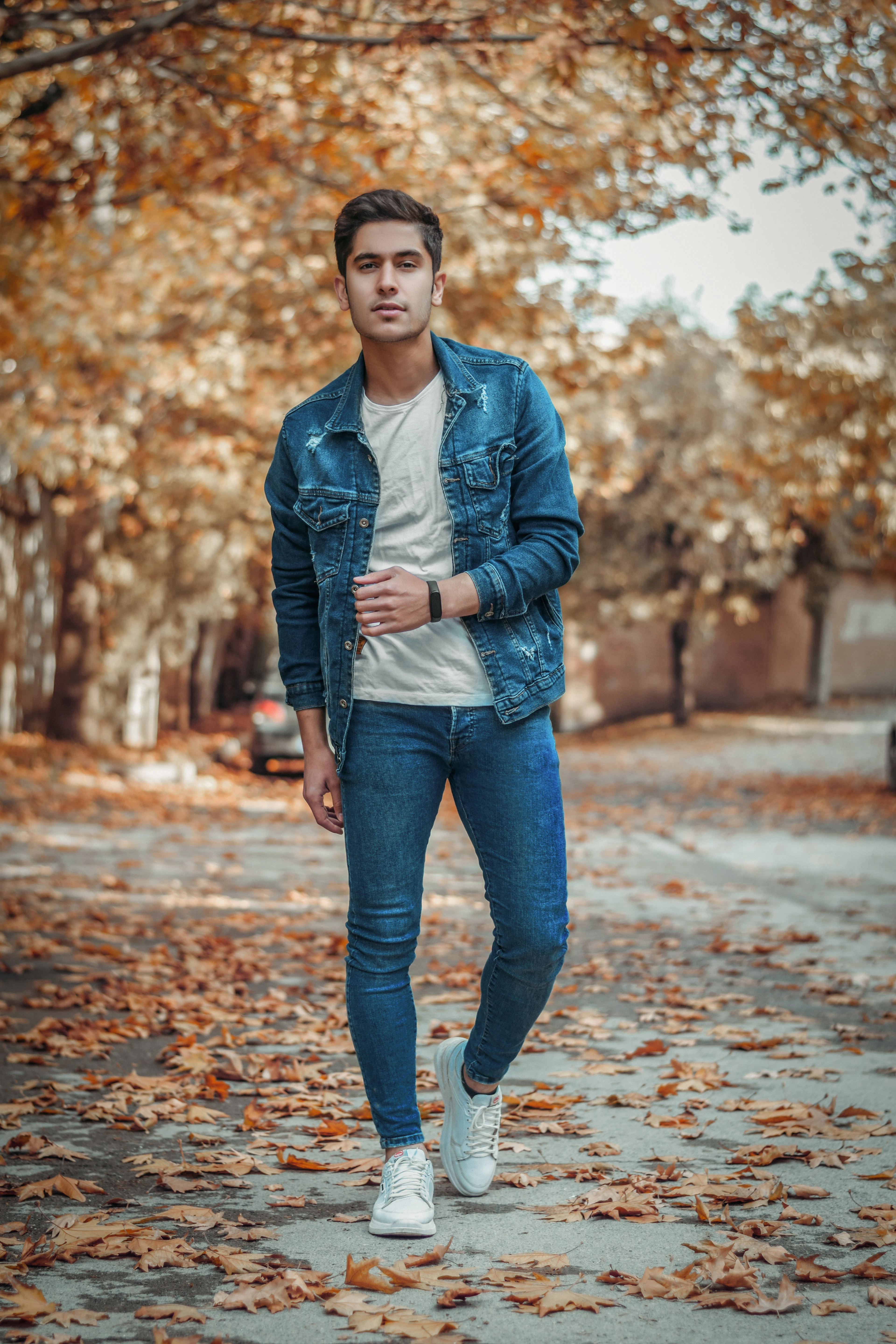 Man walking down an autumn street in a denim jacket, white tee, blue jeans and white sneakers