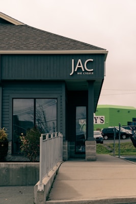 A commercial building with a modern design is visible, featuring dark gray vertical siding. The word 'JAC' and the phrase 'we create' are prominently displayed on the facade. The building has large windows and a flat, concrete pathway leading to an entrance. In the background, there is a green building and parked cars.