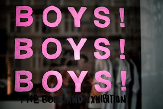 Pink letters spelling out 'BOYS!' are repeated three times vertically on a reflective surface, possibly glass. Below the repeated word, a phrase 'THE BOOK AND EXHIBITION' is written in black paint. The background shows a blurred reflection of a street scene with buildings and possibly some people.