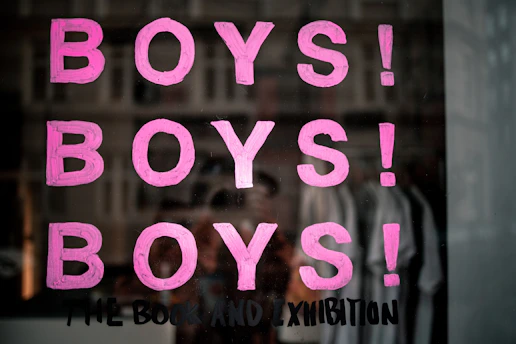 Pink letters spelling out 'BOYS!' are repeated three times vertically on a reflective surface, possibly glass. Below the repeated word, a phrase 'THE BOOK AND EXHIBITION' is written in black paint. The background shows a blurred reflection of a street scene with buildings and possibly some people.