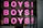 Pink letters spelling out 'BOYS!' are repeated three times vertically on a reflective surface, possibly glass. Below the repeated word, a phrase 'THE BOOK AND EXHIBITION' is written in black paint. The background shows a blurred reflection of a street scene with buildings and possibly some people.