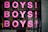Pink letters spelling out 'BOYS!' are repeated three times vertically on a reflective surface, possibly glass. Below the repeated word, a phrase 'THE BOOK AND EXHIBITION' is written in black paint. The background shows a blurred reflection of a street scene with buildings and possibly some people.