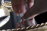 A hand holding a large, sparkling diamond between thumb and forefinger over a reflective surface, likely a mirror. The background is adorned with an ornate, golden frame featuring intricate designs.