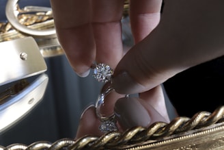 A hand holding a large, sparkling diamond between thumb and forefinger over a reflective surface, likely a mirror. The background is adorned with an ornate, golden frame featuring intricate designs.