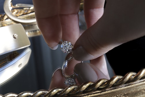 A hand holding a large, sparkling diamond between thumb and forefinger over a reflective surface, likely a mirror. The background is adorned with an ornate, golden frame featuring intricate designs.
