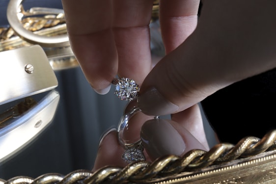 A hand holding a large, sparkling diamond between thumb and forefinger over a reflective surface, likely a mirror. The background is adorned with an ornate, golden frame featuring intricate designs.