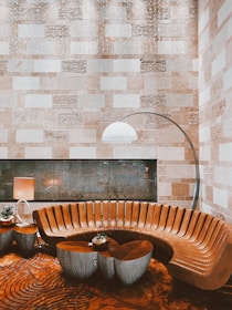 The image depicts a modern and elegant interior space featuring a semi-circular brown leather sofa with sleek, vertically-aligned panels. In front of the sofa, there are artistic, metallic side tables with intricate cut-out designs, one of which holds a small potted plant. To the left of the sofa, there is a contemporary lamp with a beige shade. A tall arc floor lamp with a white dome shade extends over the sofa, adding a stylish and functional lighting element. The background showcases a textured wall composed of alternating rectangular stone tiles in various shades of beige and cream. The floor features a bold, patterned carpet with circular and spiral designs.