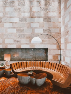The image depicts a modern and elegant interior space featuring a semi-circular brown leather sofa with sleek, vertically-aligned panels. In front of the sofa, there are artistic, metallic side tables with intricate cut-out designs, one of which holds a small potted plant. To the left of the sofa, there is a contemporary lamp with a beige shade. A tall arc floor lamp with a white dome shade extends over the sofa, adding a stylish and functional lighting element. The background showcases a textured wall composed of alternating rectangular stone tiles in various shades of beige and cream. The floor features a bold, patterned carpet with circular and spiral designs.