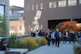 A group of people is gathered in an urban outdoor space featuring plants and trees with autumn foliage. A mural of a figure holding a sign that reads 'Love is the answer' is painted on the side of a building. The scene includes a mix of individuals either walking or standing, engaging in conversation, or using their phones, with a background of city architecture.