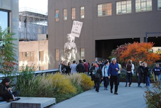 A group of people is gathered in an urban outdoor space featuring plants and trees with autumn foliage. A mural of a figure holding a sign that reads 'Love is the answer' is painted on the side of a building. The scene includes a mix of individuals either walking or standing, engaging in conversation, or using their phones, with a background of city architecture.