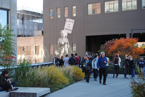 A group of people is gathered in an urban outdoor space featuring plants and trees with autumn foliage. A mural of a figure holding a sign that reads 'Love is the answer' is painted on the side of a building. The scene includes a mix of individuals either walking or standing, engaging in conversation, or using their phones, with a background of city architecture.